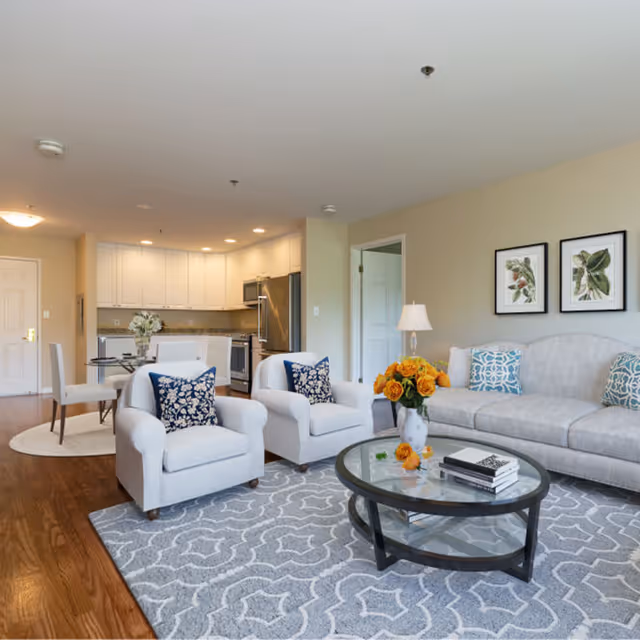 Bright open living room with a sofa, two armchairs, a round glass coffee table with flowers, and an open kitchen in the background.