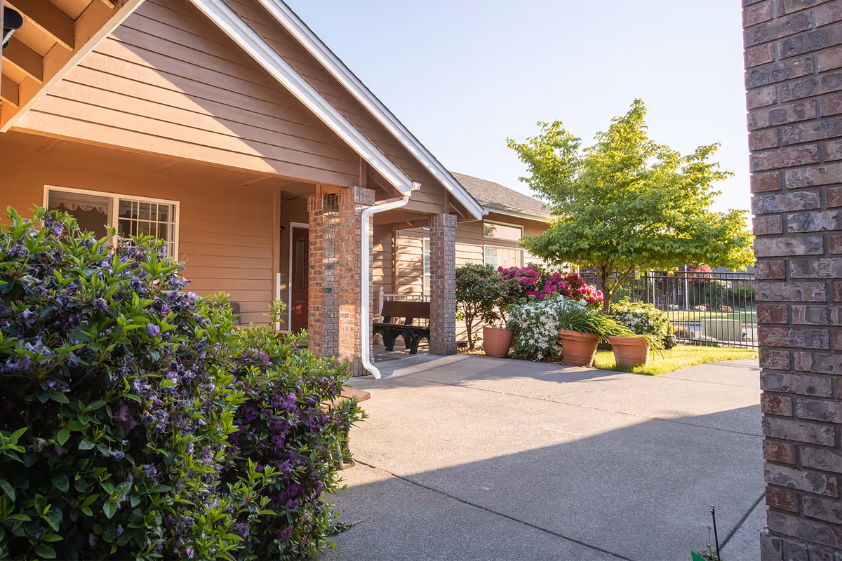 Outdoor view of a senior living facility with a concrete pathway, brown siding building with a covered porch supported by brick columns, potted plants with colorful flowers, green shrubs, and a tree under a clear sky.