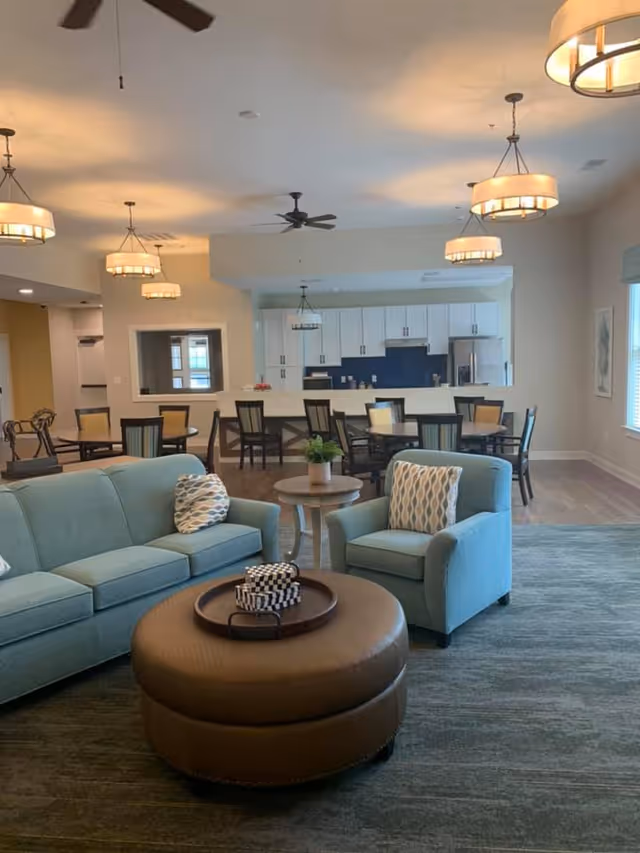 A spacious living area with a light blue sofa and matching armchair, a round brown ottoman with a tray on top, and a small round side table with a plant. In the background, there is a dining area with multiple tables and chairs, and a kitchen with white cabinets and a blue backsplash. The room is well-lit with ceiling lights and ceiling fans.