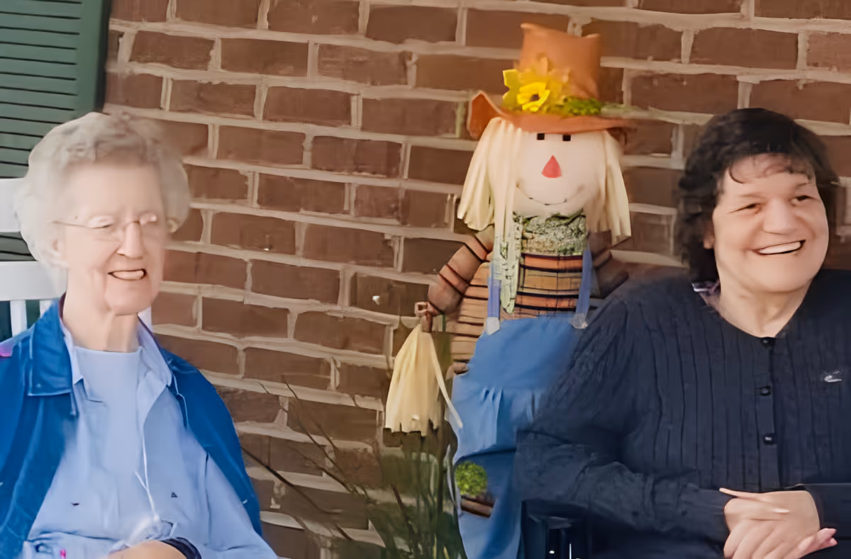 Two elderly women sitting and smiling on a porch with a brick wall background. Between them is a decorative scarecrow figure wearing a hat with a sunflower and blue overalls.