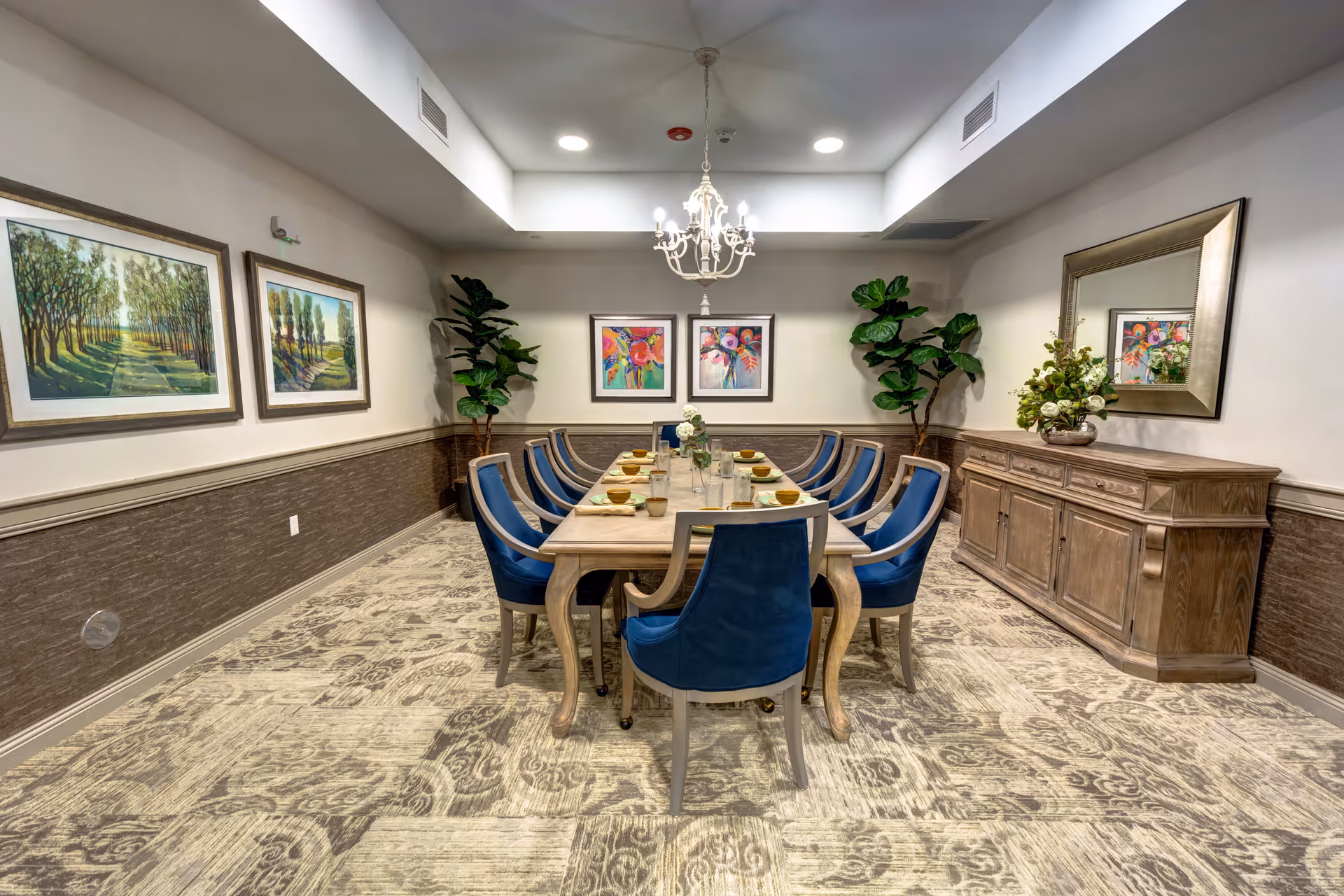 A well-decorated dining room with a long wooden table surrounded by blue upholstered chairs. The table is set with plates, glasses, and napkins. The room features a chandelier hanging from the ceiling, framed artwork on the walls, two large potted plants in the corners, a wooden sideboard with a flower arrangement, and a large mirror above it. The floor has a patterned carpet.