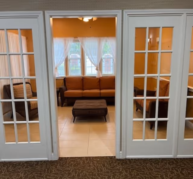 A cozy living room area viewed through white French doors with glass panes. Inside, there is a brown sofa, two matching armchairs, and a woven ottoman in the center. The room has beige tiled flooring and three windows with sheer white curtains letting in natural light.