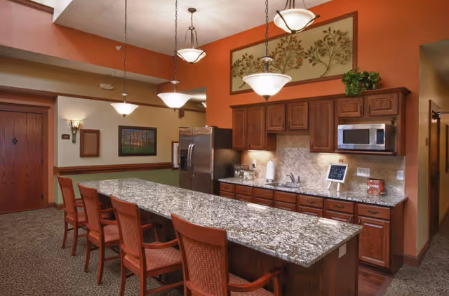 Communal kitchen area with a long granite island, wooden chairs, stainless steel refrigerator, cabinets and hanging pendant lights.