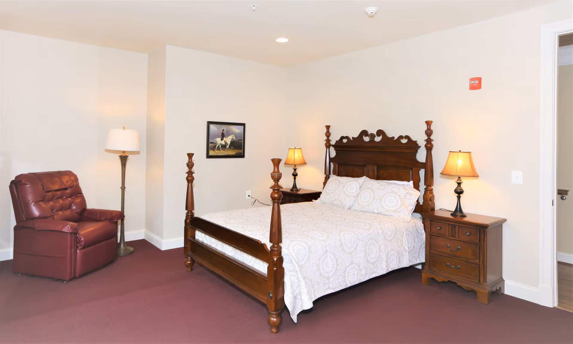 A bedroom with a wooden four-poster bed covered with a white patterned bedspread and two matching pillows. On either side of the bed are wooden nightstands with table lamps. To the left of the bed is a maroon leather recliner chair and a floor lamp. A framed picture hangs on the wall above the bed. The room has beige walls and a maroon carpet.