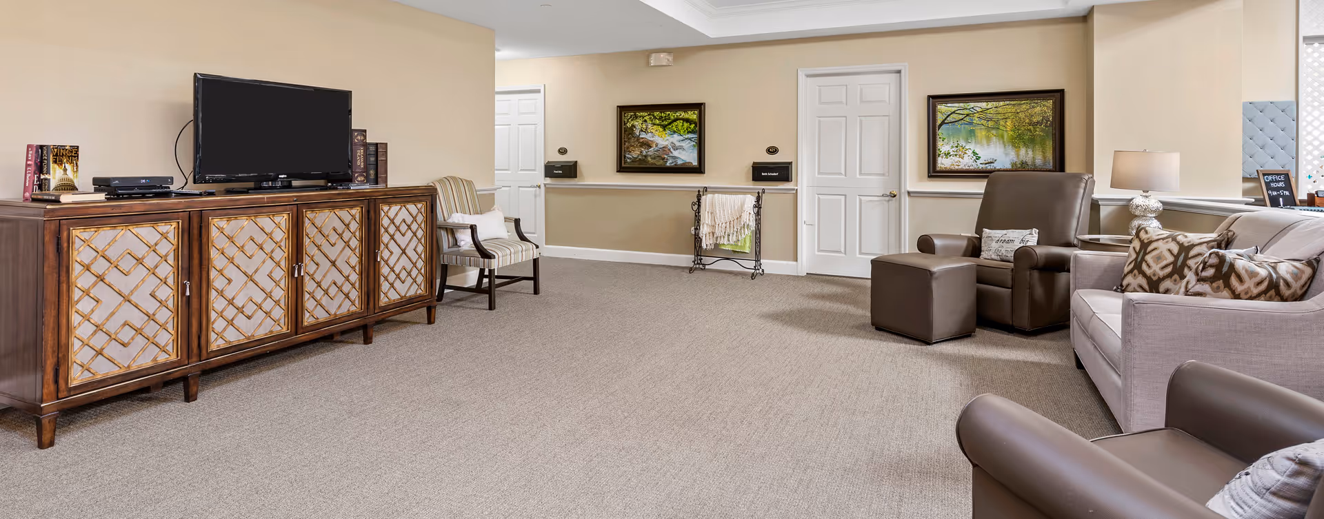 A spacious living room area with beige walls and carpeted floor. The room features a wooden cabinet with a flat-screen TV on top, a striped armchair, a brown leather recliner with an ottoman, a light gray sofa with patterned pillows, and a small table lamp. Two framed landscape paintings hang on the walls, and there is a small rack with folded blankets near a white door.