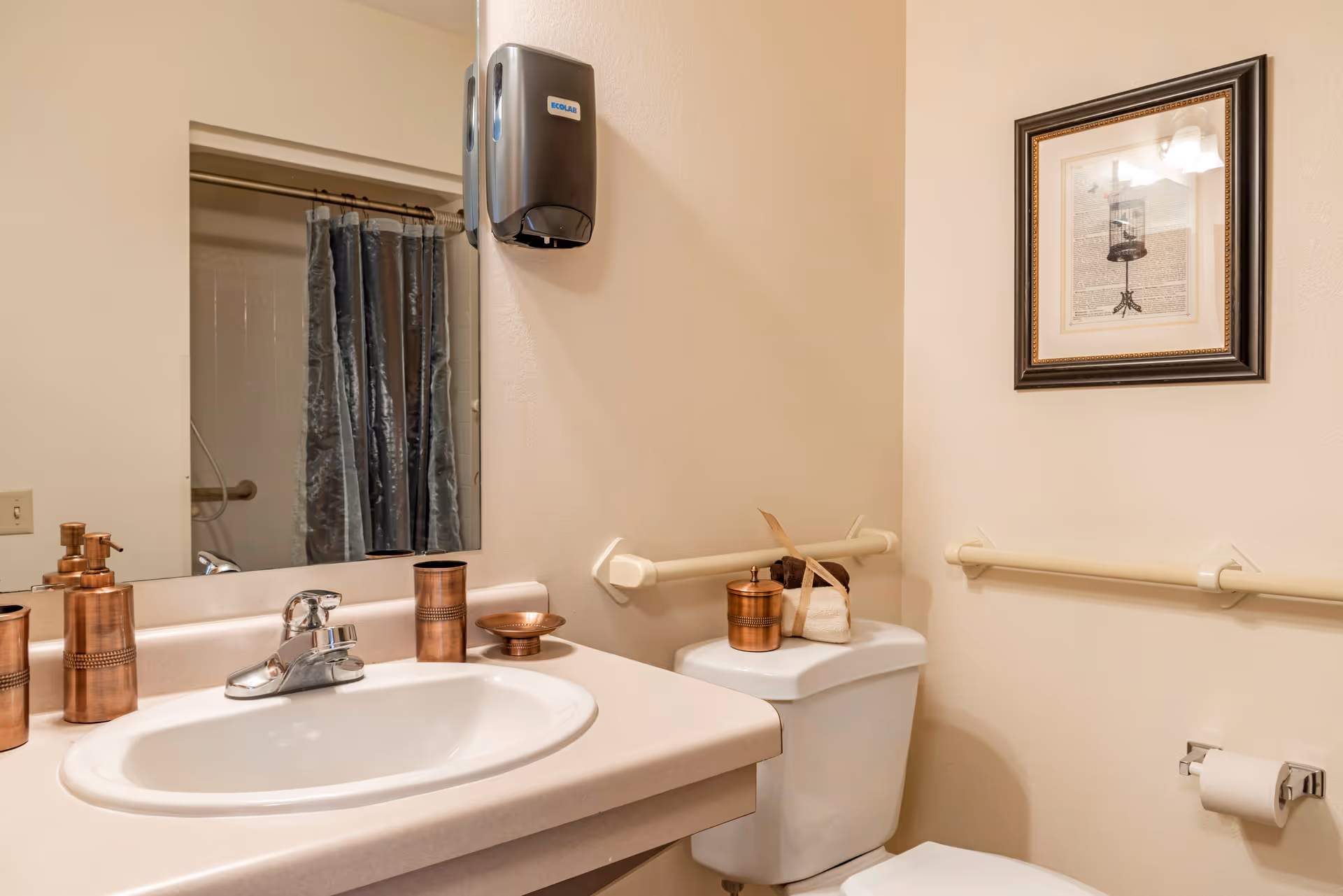 A bathroom with a white sink and countertop, a silver faucet, and copper-colored soap dispenser, cup, and dish. A toilet with a copper container and folded towels on top is next to the sink. Beige walls have safety grab bars and a framed picture. A shower with a gray curtain is visible in the mirror reflection.