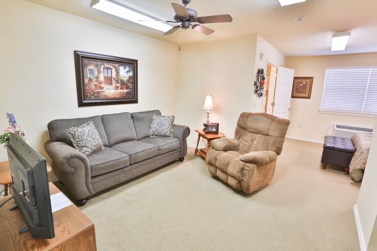 A cozy living room area in Wesley House featuring a gray upholstered sofa with two patterned cushions, a brown recliner chair, a wooden side table with a lamp and framed photo, a flat-screen TV on a wooden stand, a ceiling fan, and a framed painting on the wall. The room has beige carpet and walls, with a window covered by blinds and a partially visible bed and ottoman in the background.