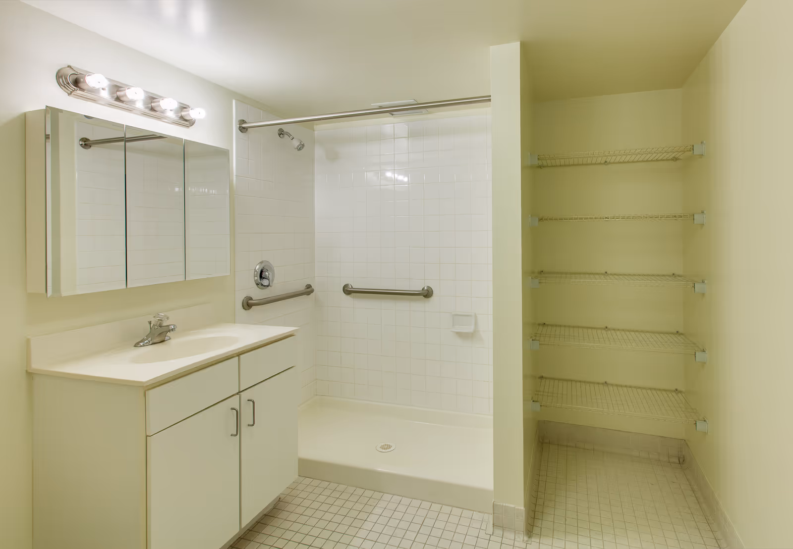 A clean bathroom featuring a white vanity with a sink and a mirrored medicine cabinet above it. Next to the vanity is a walk-in shower with white tiled walls, equipped with two grab bars and a showerhead. To the right of the shower, there is an open shelving unit with five wire shelves. The floor is covered with small square tiles.