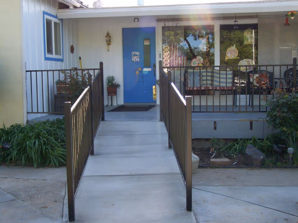 Front entrance of a care home with a blue door, a concrete ramp with black metal railings on both sides, and a porch area with a large window decorated with hanging ornaments and a striped blanket on a chair.