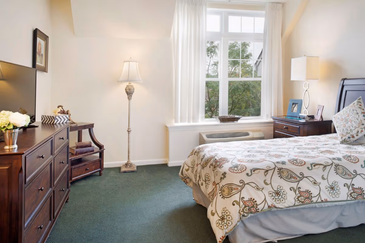 Sunlit bedroom with a patterned bed, wooden dressers, lamps, and a large window overlooking trees.