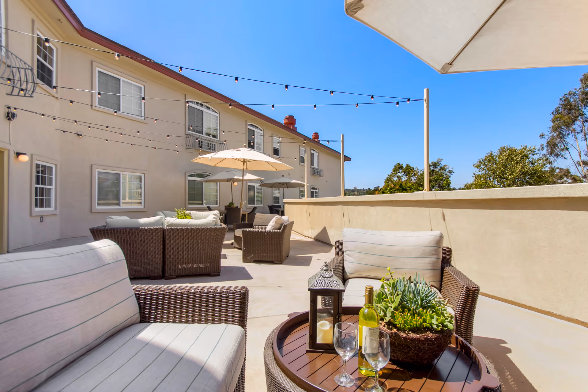 Outdoor patio area at a senior living facility with wicker furniture including cushioned chairs and sofas, a round table with a bottle of white wine, two wine glasses, a lantern, and a potted succulent plant. There are string lights overhead and umbrellas providing shade. The building exterior with multiple windows is visible under a clear blue sky.