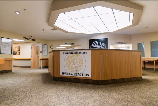 Interior view of a reception or nurse station area in a senior living facility with a large wooden counter displaying a sign that reads '#kindnessmatters SPARK a REACTION'. The ceiling has a large rectangular light panel, and there are signs on the walls indicating directions to therapy, chestnut lane, and ambulance entrance. The floor is carpeted and the walls are light-colored with framed pictures.