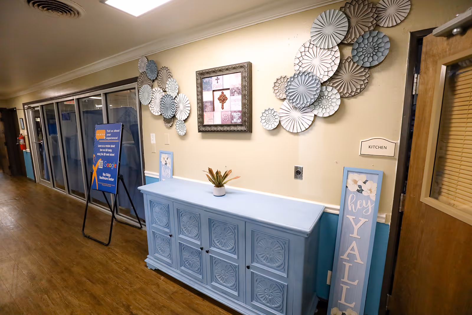 Interior hallway of Bay Ridge Healthcare Center with wooden flooring and beige walls. A light blue cabinet with decorative patterns is against the wall, topped with a small potted plant. Above the cabinet is a framed artwork and decorative circular wall hangings in shades of white, beige, and blue. To the right, a door labeled 'KITCHEN' is partially visible. A signboard with information about Bay Ridge Healthcare Center is placed along the hallway near glass doors.