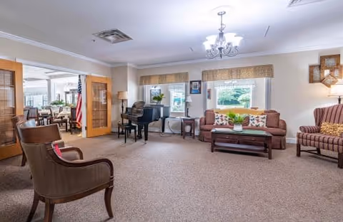 A spacious living room area in a senior living facility with a brown sofa, two armchairs, a coffee table, a piano, and large windows with beige valances letting in natural light. There is a chandelier hanging from the ceiling and an American flag visible through open double doors leading to another room.