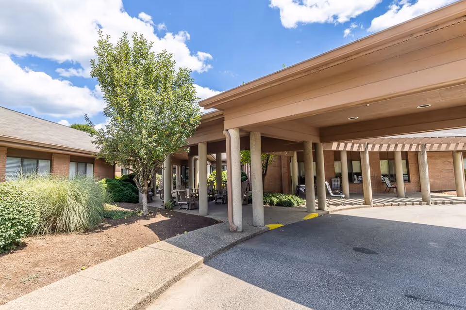 Exterior view of Elison Assisted Living & Memory Care of Marietta showing a covered entrance with columns, outdoor seating, landscaping with bushes and a tree, and a partly cloudy sky.