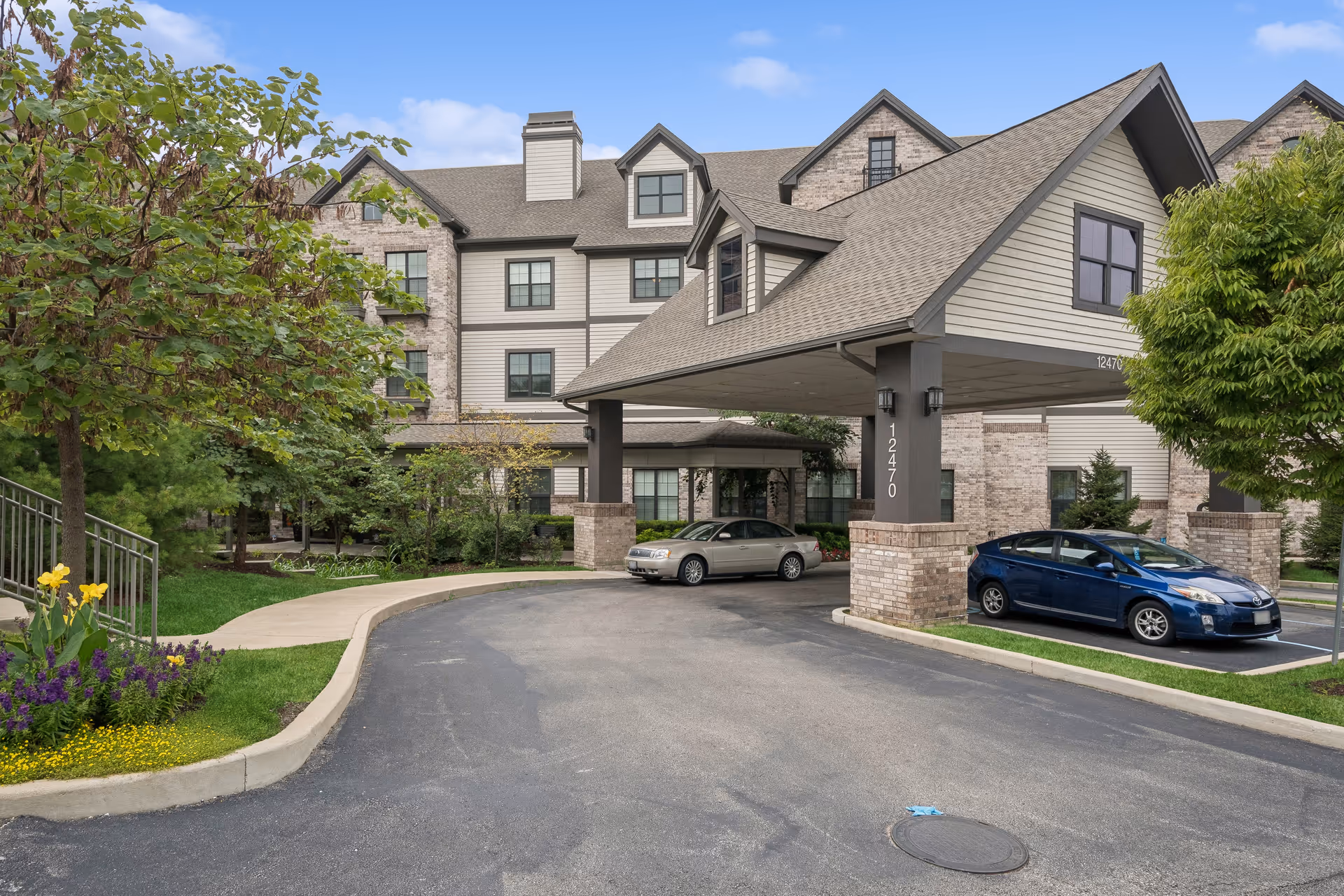 Exterior view of The Grande at Laumeier Park senior living facility showing the front entrance with a covered drop-off area. Two cars are parked near the entrance, surrounded by landscaped greenery and trees under a blue sky with some clouds.