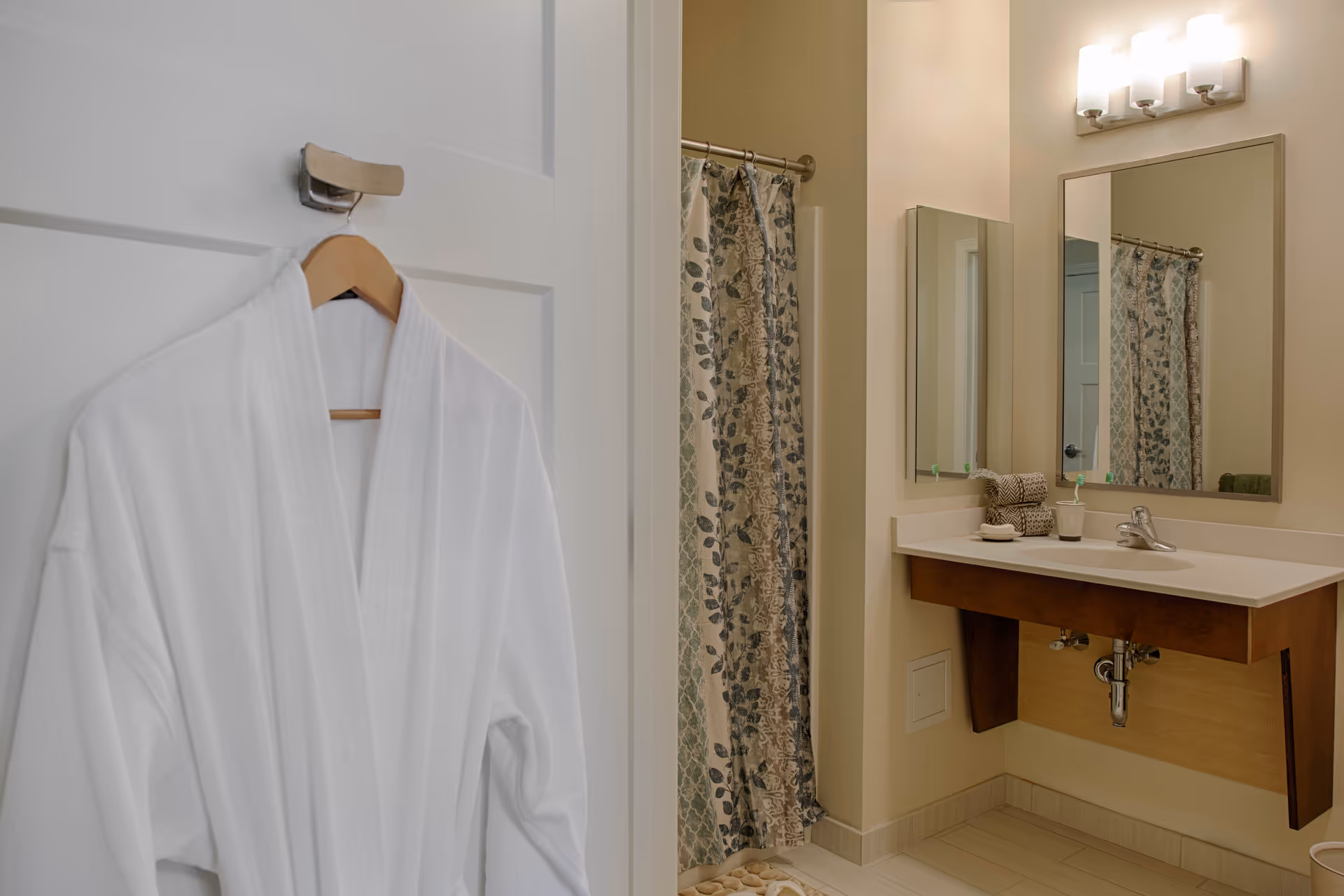 A bathroom interior featuring a white bathrobe hanging on a door hook, a shower area with a patterned curtain, a sink with a wooden vanity, a large mirror, and a light fixture above the mirror.