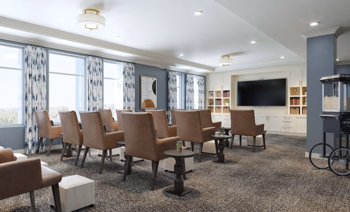 A spacious, well-lit common area with rows of brown upholstered chairs facing a large flat-screen TV mounted on a white built-in shelving unit. The room has large windows with patterned curtains allowing natural light to fill the space, a popcorn machine on the right side, and small side tables next to some chairs. The carpet is a textured gray, and the walls are painted in a soft blue-gray tone.