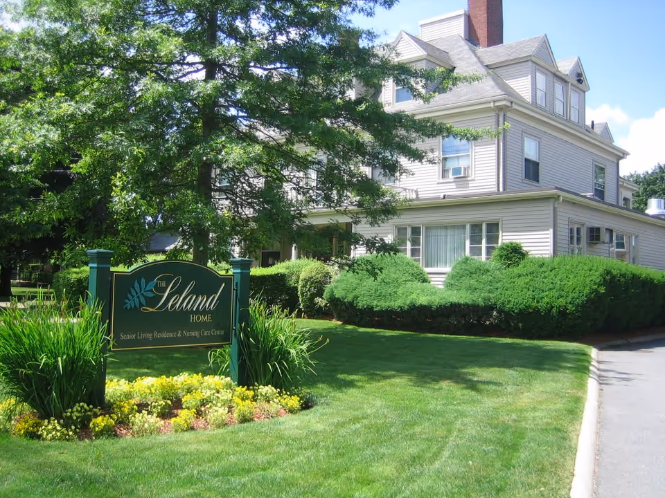 Multi-story house-like senior living building with manicured lawn and a front sign reading 'The Leland Home'.