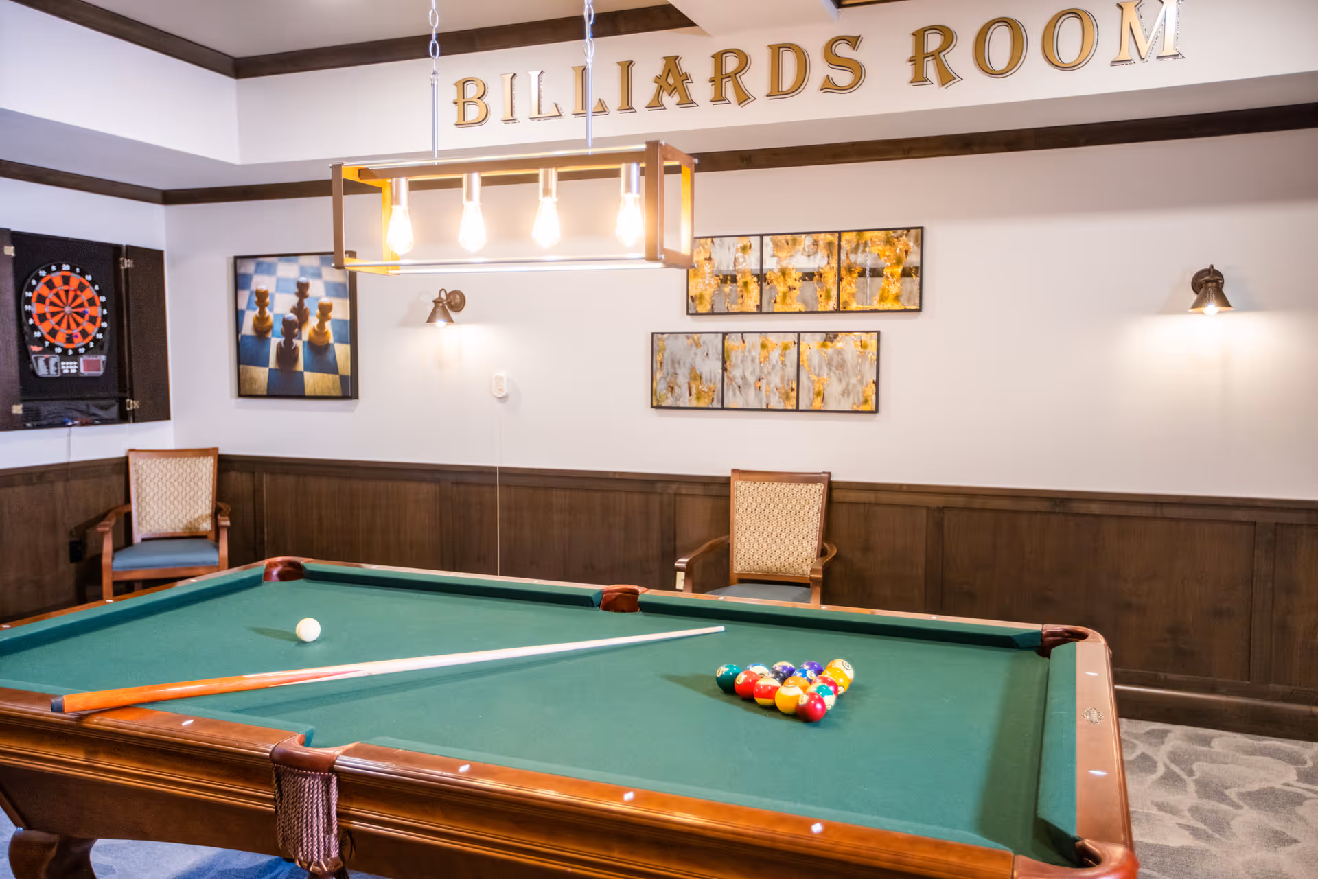 Interior view of a billiards room with a pool table in the center, a cue stick and billiard balls arranged on the table. The room has wooden paneling on the walls, two chairs, a dartboard cabinet, and framed artwork. A light fixture with exposed bulbs hangs above the pool table, and the words 'BILLIARDS ROOM' are displayed on the wall above.
