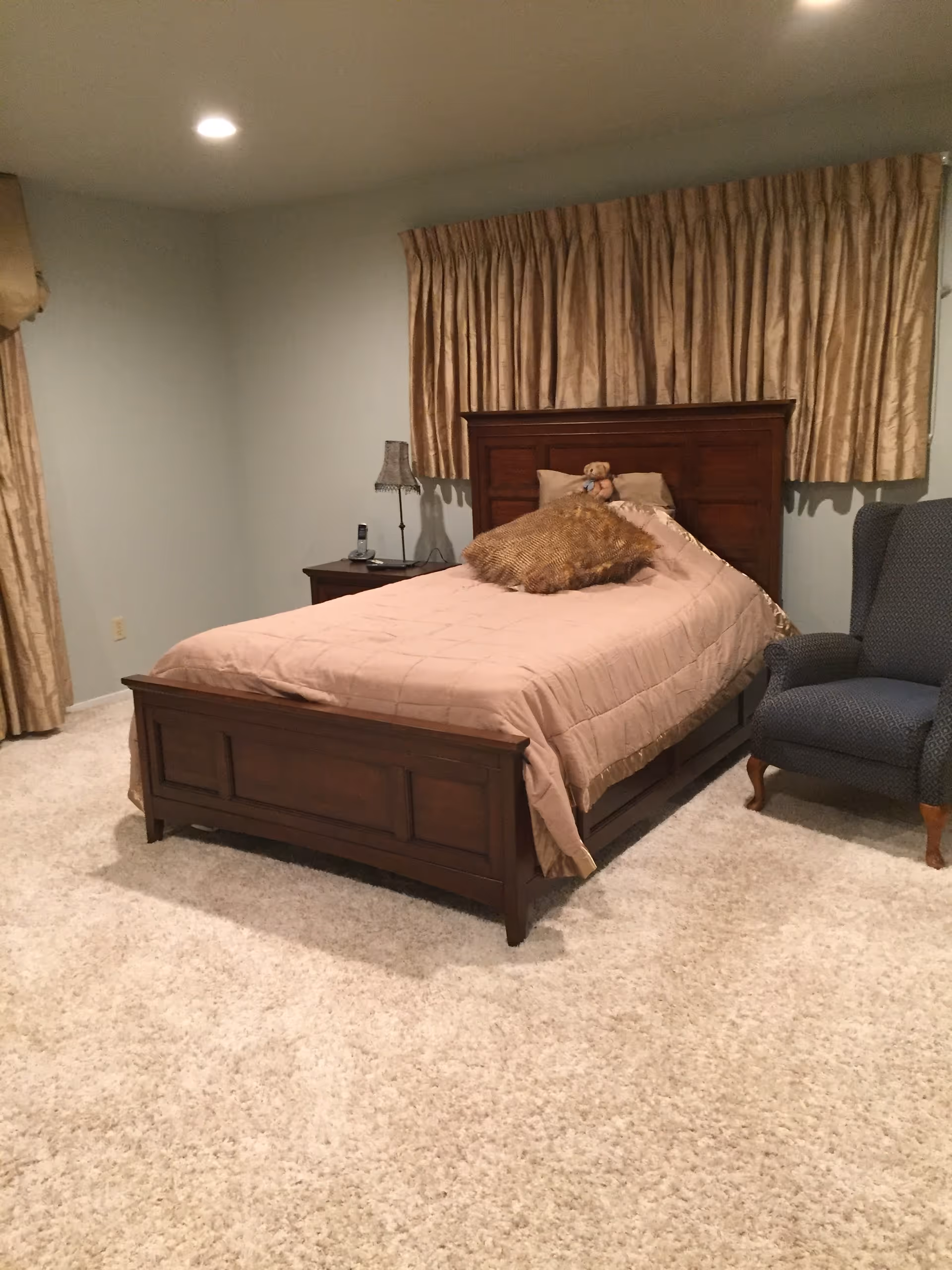 A cozy bedroom with a wooden single bed dressed in beige bedding, a nightstand with a lamp, and an upholstered armchair on a carpeted floor.