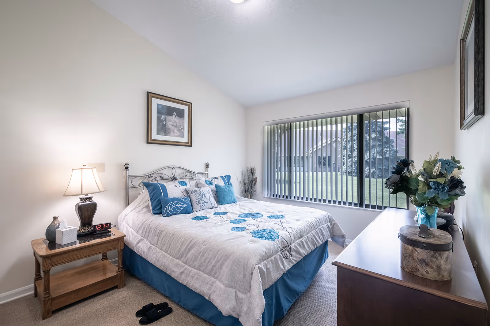 A bright furnished bedroom with a bed, nightstand, dresser and a large window with vertical blinds.