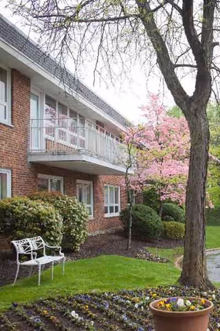 Brick senior living building exterior with balconies, trees, bench, and flower garden.