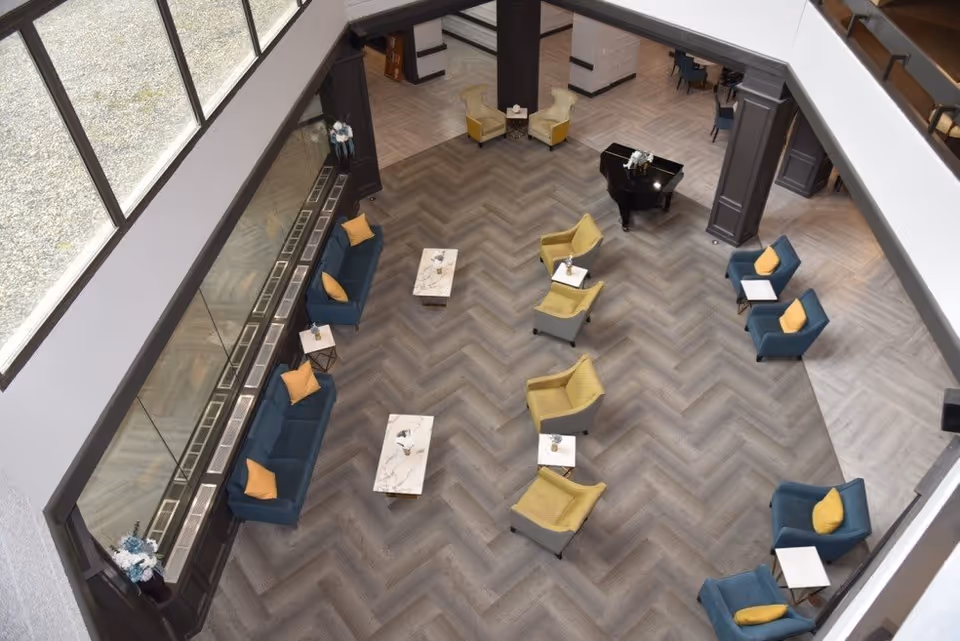 Overhead view of a spacious senior living facility common area with a herringbone patterned floor. The room features multiple seating arrangements including blue sofas with yellow pillows, yellow armchairs, small white side tables, and a black grand piano. Large windows on one side allow natural light to fill the space.
