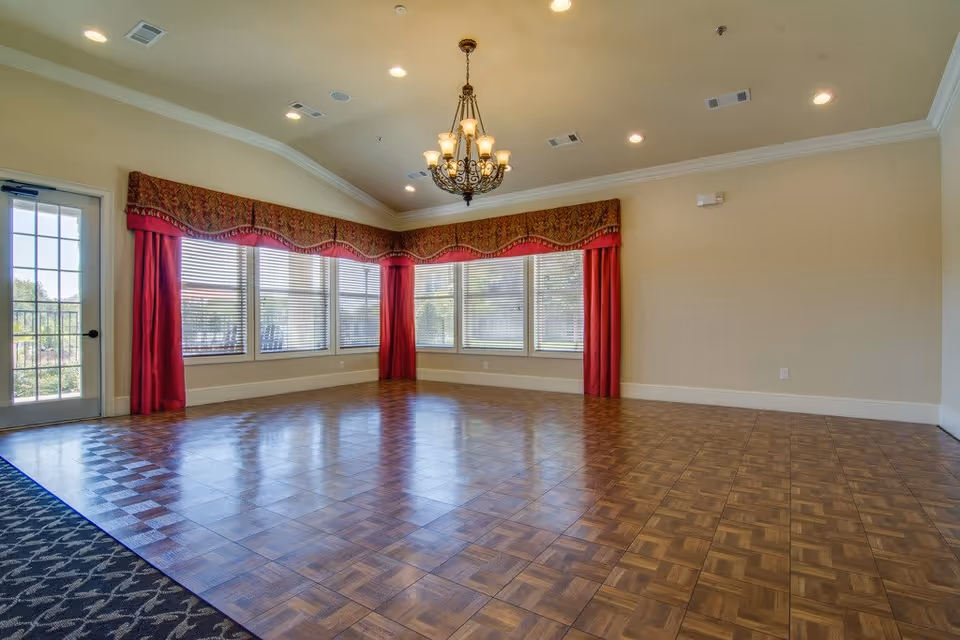 Empty community room with parquet flooring, a chandelier, and large windows with red valances and curtains.