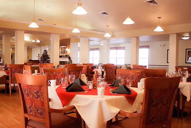 A dining room in a senior living facility decorated for the holidays with tables covered in white tablecloths and red runners, set with glasses, cups, and black napkins. There is a Christmas tree in the background and warm lighting from ceiling lamps.