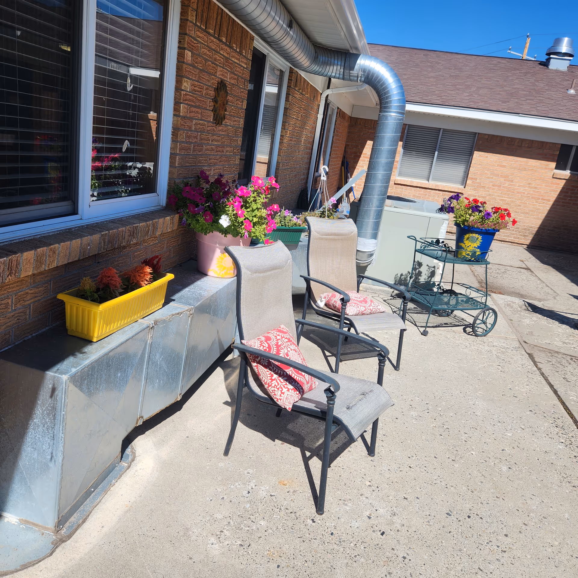 Outdoor patio area with two metal chairs featuring red patterned cushions, several flower pots with colorful flowers placed on a ledge and a small metal cart, all set against a brick building wall under a clear blue sky.
