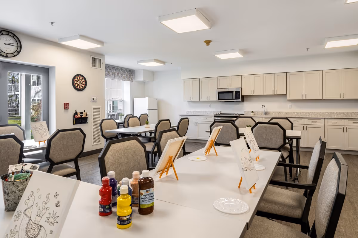 Bright activity/dining room with tables, chairs, art supplies on easels, and a kitchenette in the background.