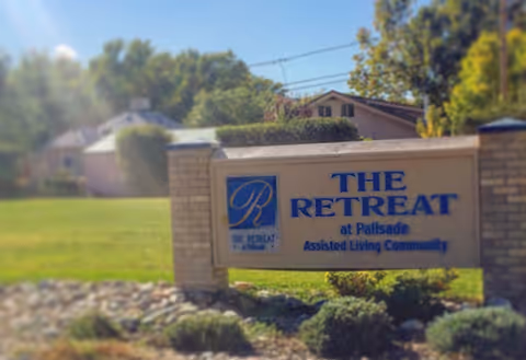 Entrance sign reading 'The Retreat at Palisade Assisted Living Community' on a stone base set on a lawn with houses and trees behind it.