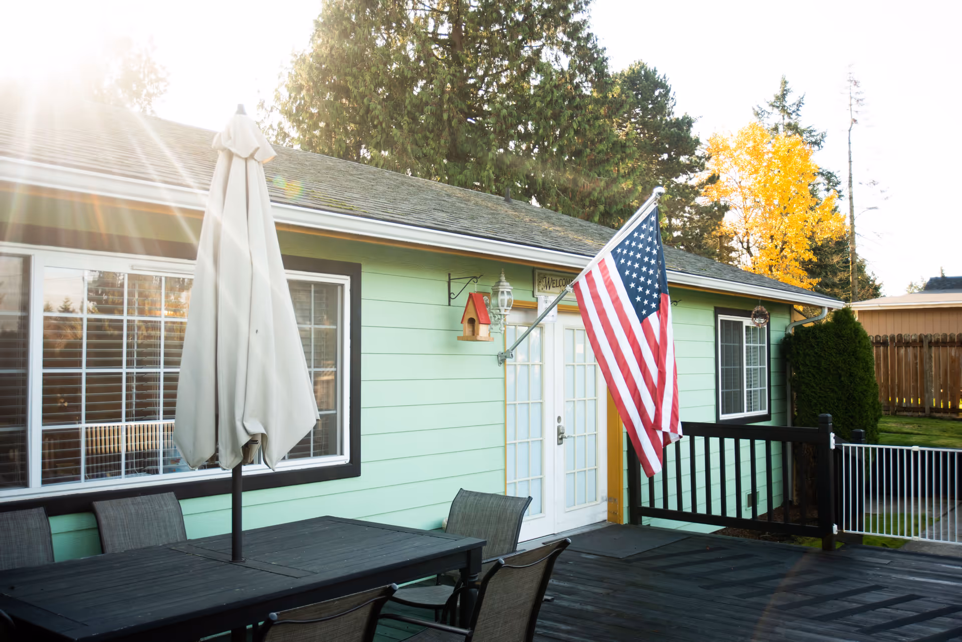 Outdoor patio area of a facility with a black wooden table and chairs, a closed beige umbrella, an American flag mounted on the light green building wall, and trees in the background with sunlight shining through.