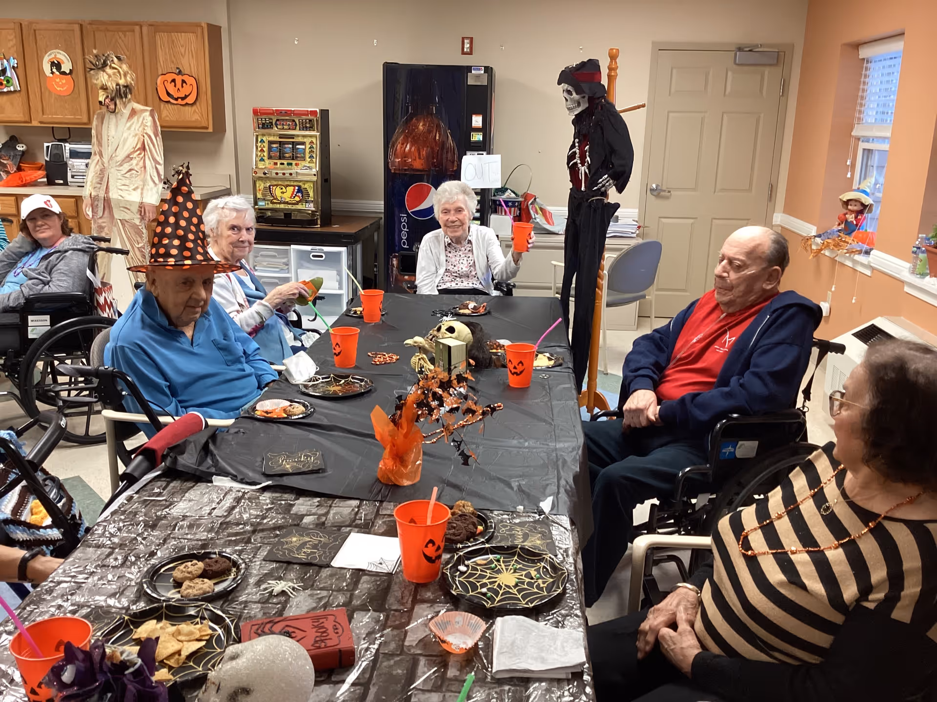 A group of elderly people sitting around a table decorated with Halloween-themed items including orange cups with jack-o'-lantern faces, spider web plates, and small decorations. Some individuals are in wheelchairs, and one person is wearing a witch hat. The room has a vending machine, cabinets with Halloween decorations, and a skeleton figure dressed as a pirate standing near the door.