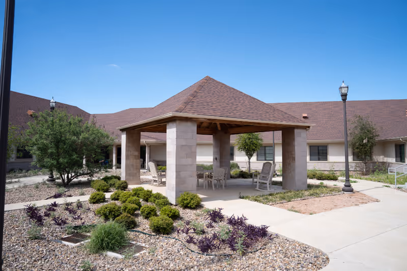 Outdoor courtyard area at Las Alturas Nursing & Transitional Care featuring a covered pavilion with a pitched roof supported by stone columns. Under the pavilion are several white chairs and a table. Surrounding the pavilion are landscaped areas with small bushes, purple and green plants, and gravel. The building with a brown shingled roof and windows is visible in the background under a clear blue sky.