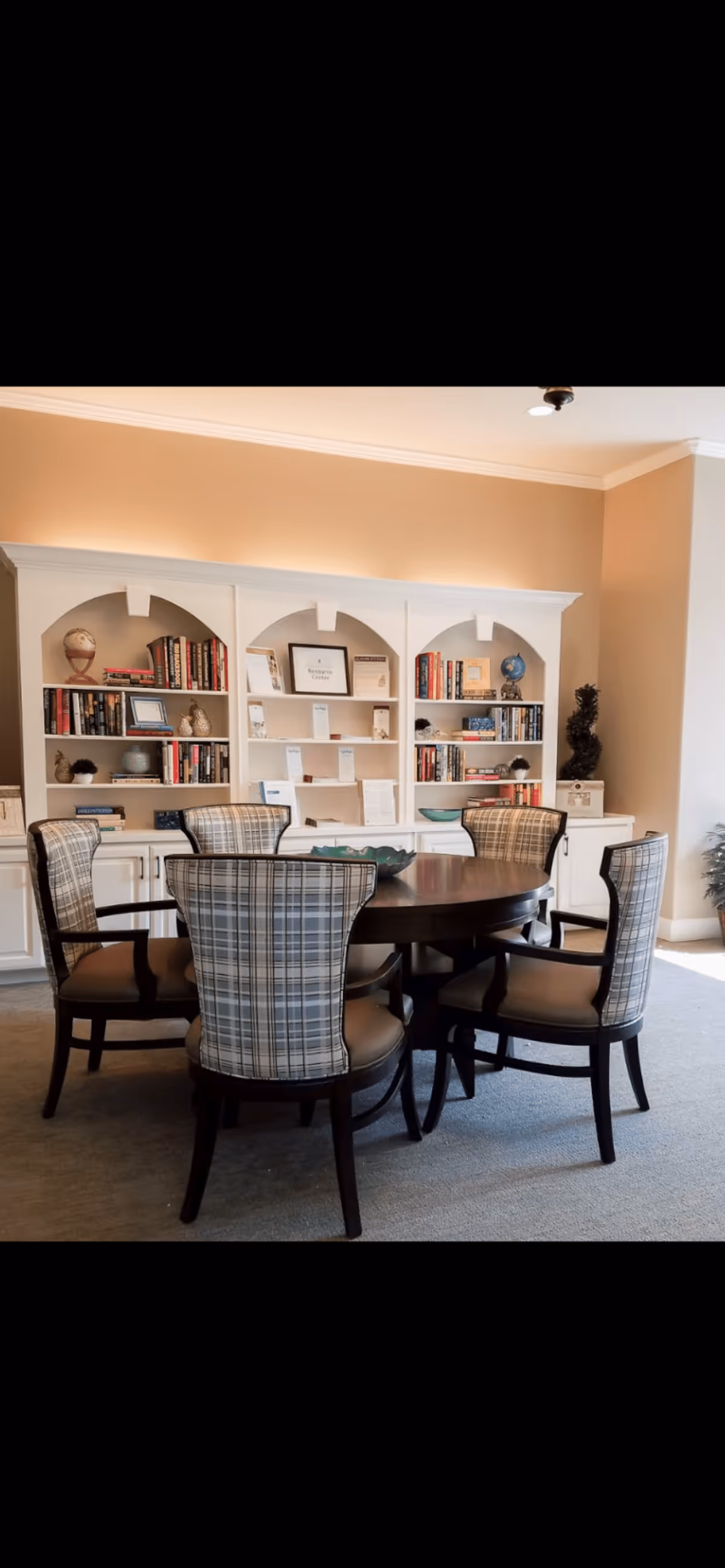 A cozy interior room featuring a round wooden table surrounded by six cushioned chairs with plaid upholstery. Behind the table is a white built-in bookshelf filled with books, decorative items, and framed documents. The room has beige walls and carpeted flooring, with soft lighting creating a warm atmosphere.