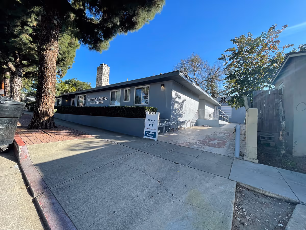 Exterior view of Whittier Pacific Care Center building on a sunny day with clear blue sky. The building is single-story with gray walls and several windows. There is a sign on the building and a sandwich board sign on the sidewalk that reads 'Whittier Pacific Welcomes You Candidate Parking'. Trees and a sidewalk are visible in the foreground.