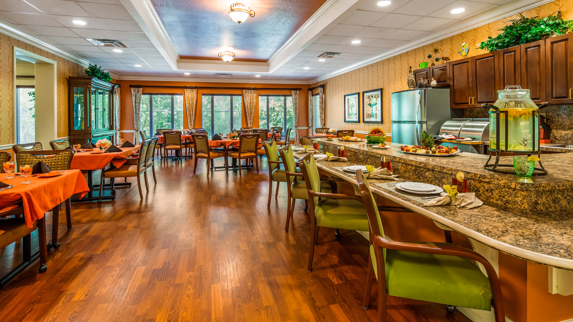 A spacious dining room with wooden flooring, multiple tables covered with orange tablecloths and set with glasses and napkins. There is a long granite counter with green cushioned chairs, plates, and a large glass dispenser filled with lemon water. The room has large windows with curtains allowing natural light to enter, and wooden cabinets and a refrigerator are visible in the background.