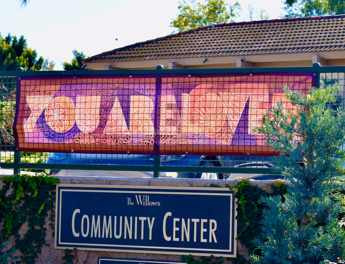 Outdoor view of a community center sign for The Willows with a colorful banner above it that reads 'YOUNG LOVE'. The scene includes greenery and a building with a tiled roof in the background.