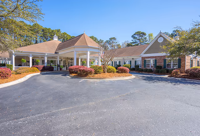 Front exterior view of Oaks At Salem Road facility showing a single-story building with a covered entrance, landscaped bushes, and a circular driveway under a clear blue sky.