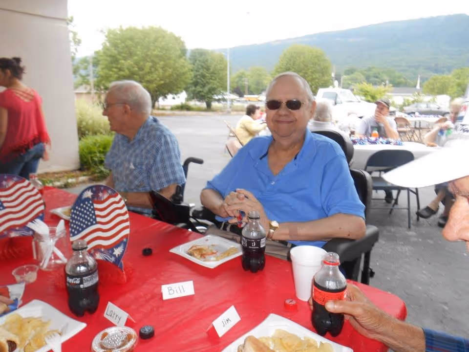 A group of elderly people sitting outdoors at a table covered with a red tablecloth decorated with American flag-themed centerpieces. They are enjoying a meal with plates of food and bottles of soda. One man in a blue shirt and sunglasses is smiling at the camera. Trees and hills are visible in the background.