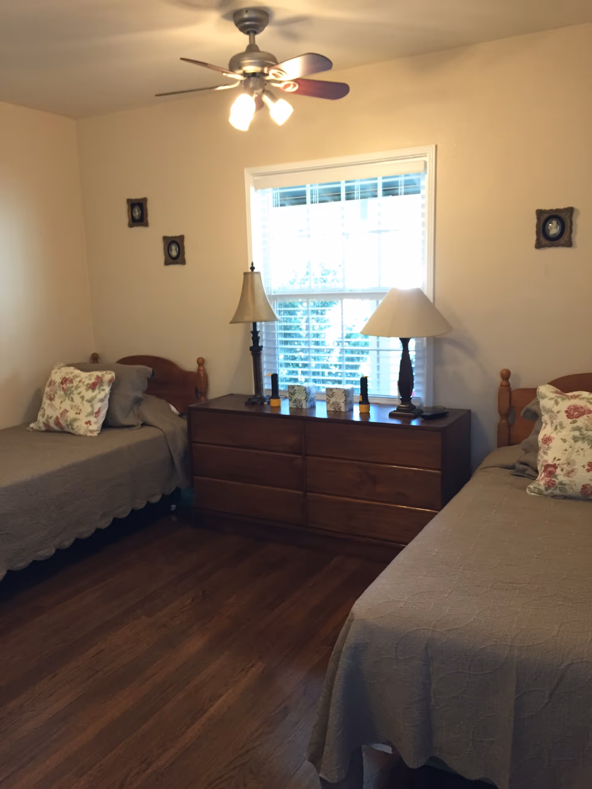 A bedroom with two twin beds, a wooden dresser beneath a window, two table lamps, and a ceiling fan.