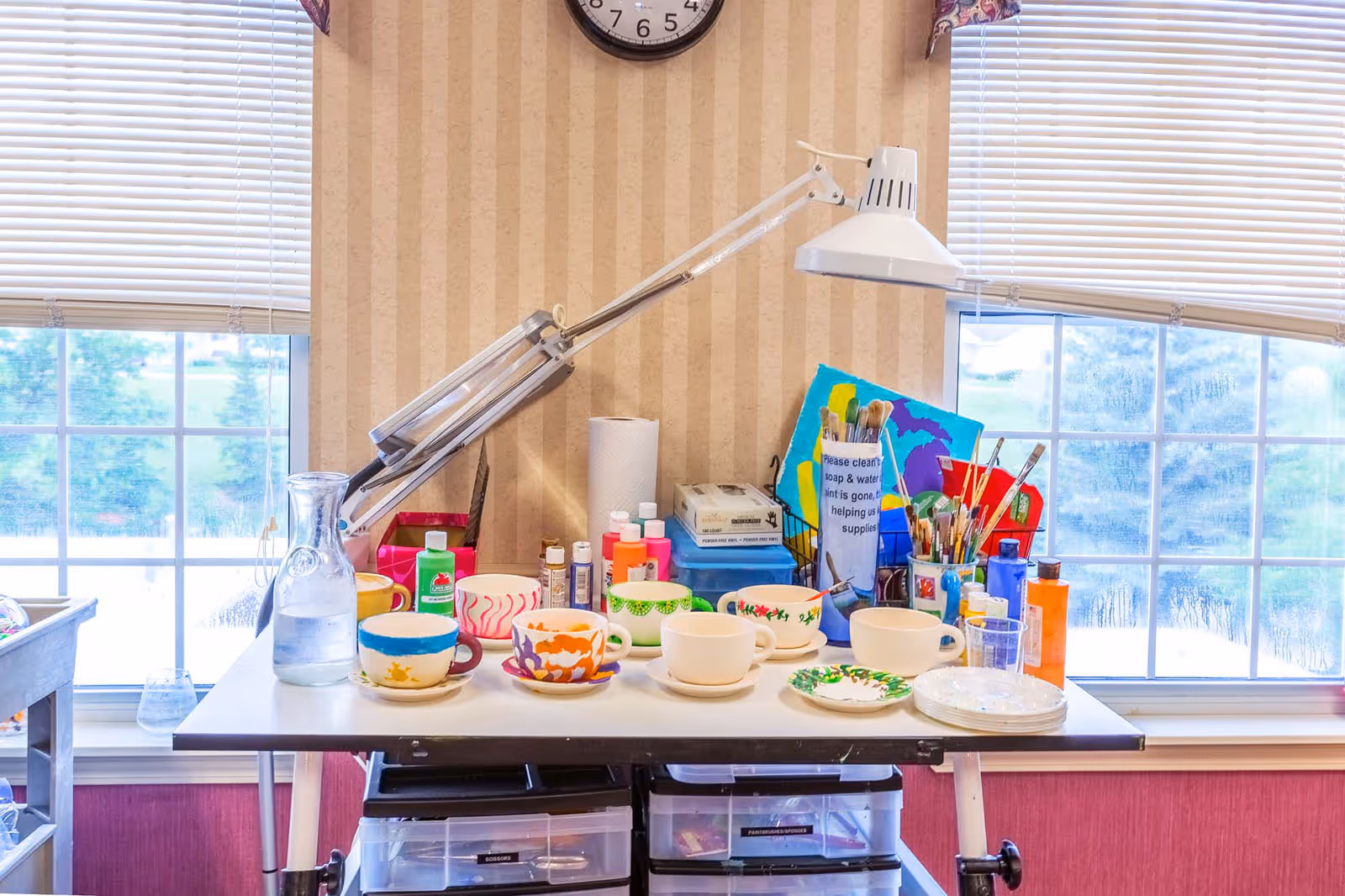 A bright art and craft workspace with a white table holding several colorful painted cups and saucers, paint bottles, paintbrushes in containers, a glass water pitcher, and a white adjustable desk lamp. The background features two large windows with white blinds and a beige striped wallpaper with a clock above the table.