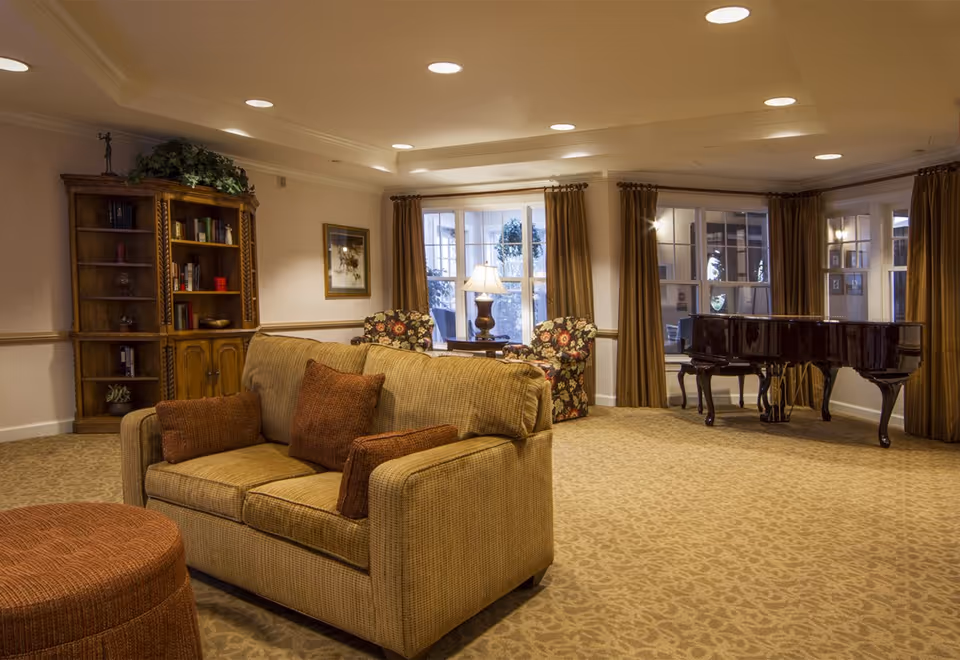 A cozy living room with a beige sofa adorned with brown cushions, a round ottoman, two floral armchairs, a wooden bookshelf with books and decorative items, a table lamp on a side table, large windows with brown curtains, and a black grand piano in the corner.