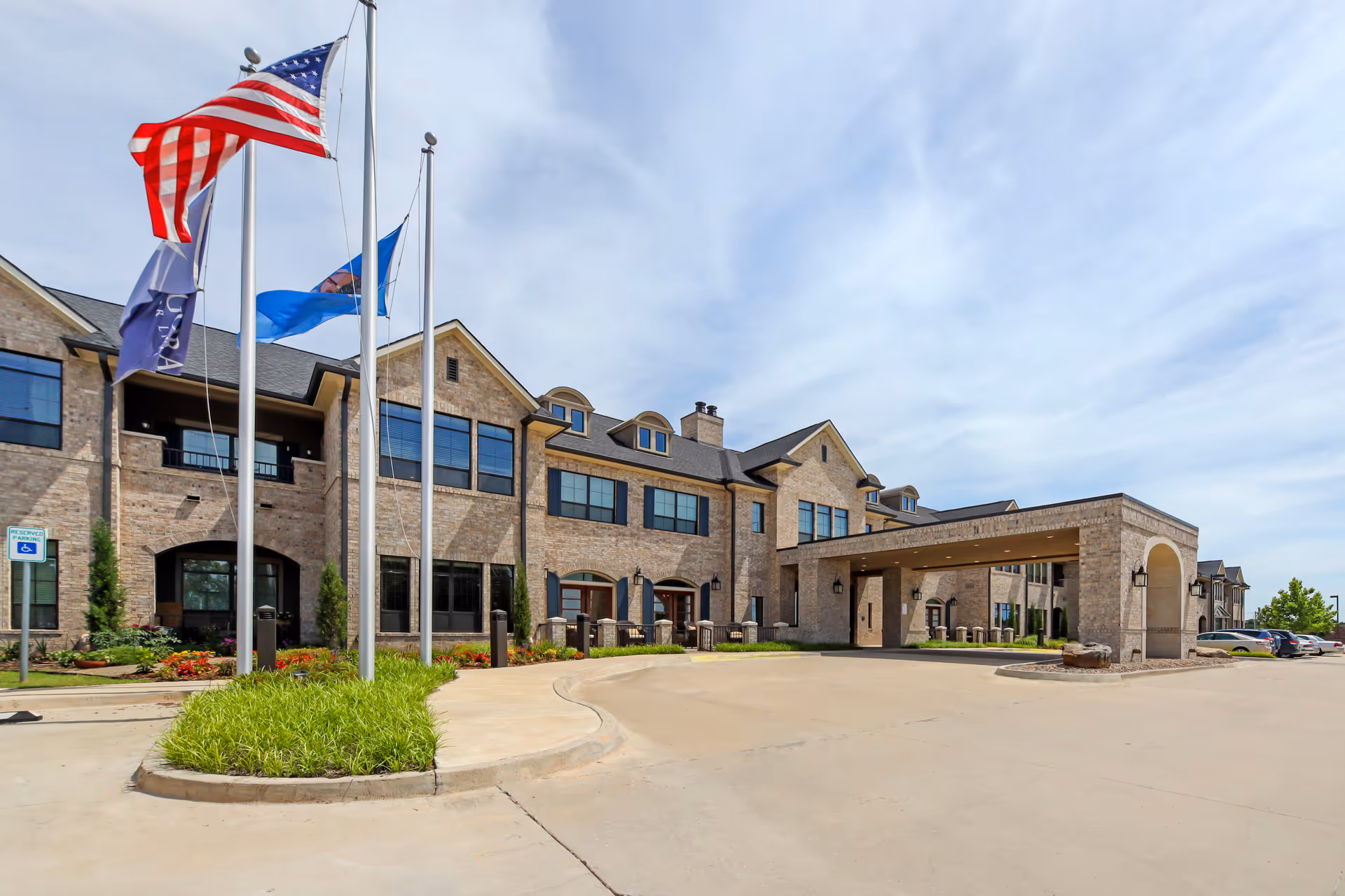 Exterior view of a large senior living facility building with beige brick walls, multiple windows, and a covered entrance. Three flagpoles with flags, including the American flag, stand in front of the building near a landscaped area with green plants and flowers. A parking lot with cars is visible on the right side under a partly cloudy sky.