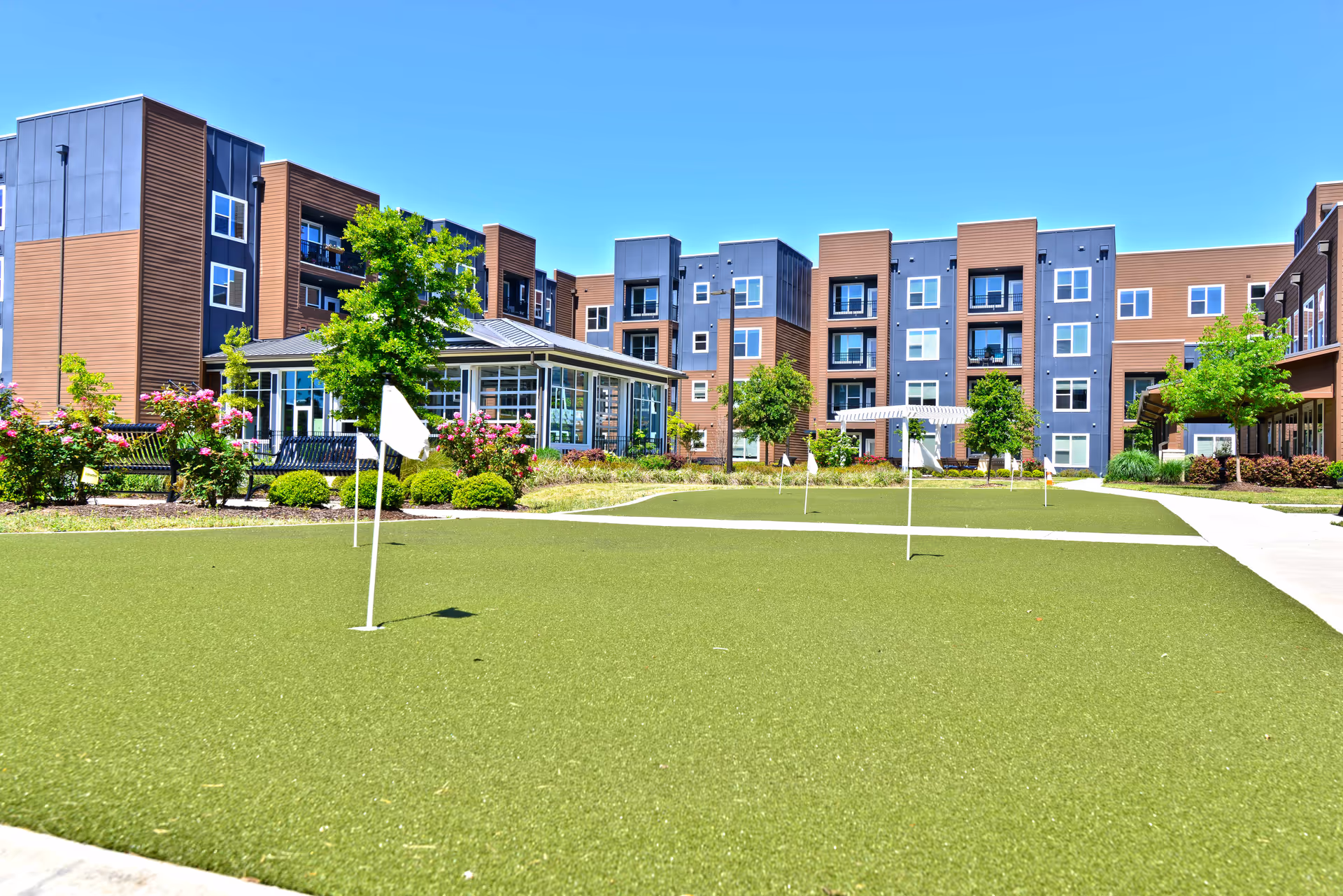 Courtyard putting green with flag markers in front of a multi-story senior living building and landscaped gardens.