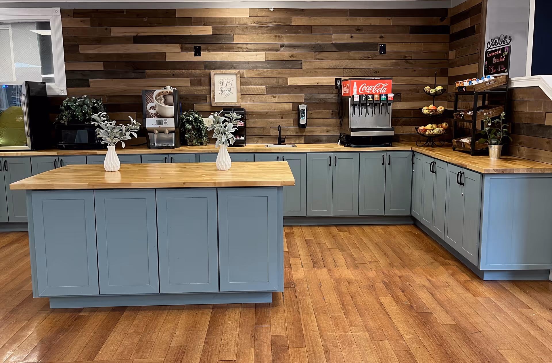 A modern kitchen area with light blue cabinets and wooden countertops. There is a central island with two small white vases holding green plants. The back wall is covered with horizontal wooden panels. On the countertop, there is a coffee machine, a Coca-Cola soda dispenser, a hand sanitizer dispenser, and a tiered fruit basket with apples. A small chalkboard sign is visible on the right side.
