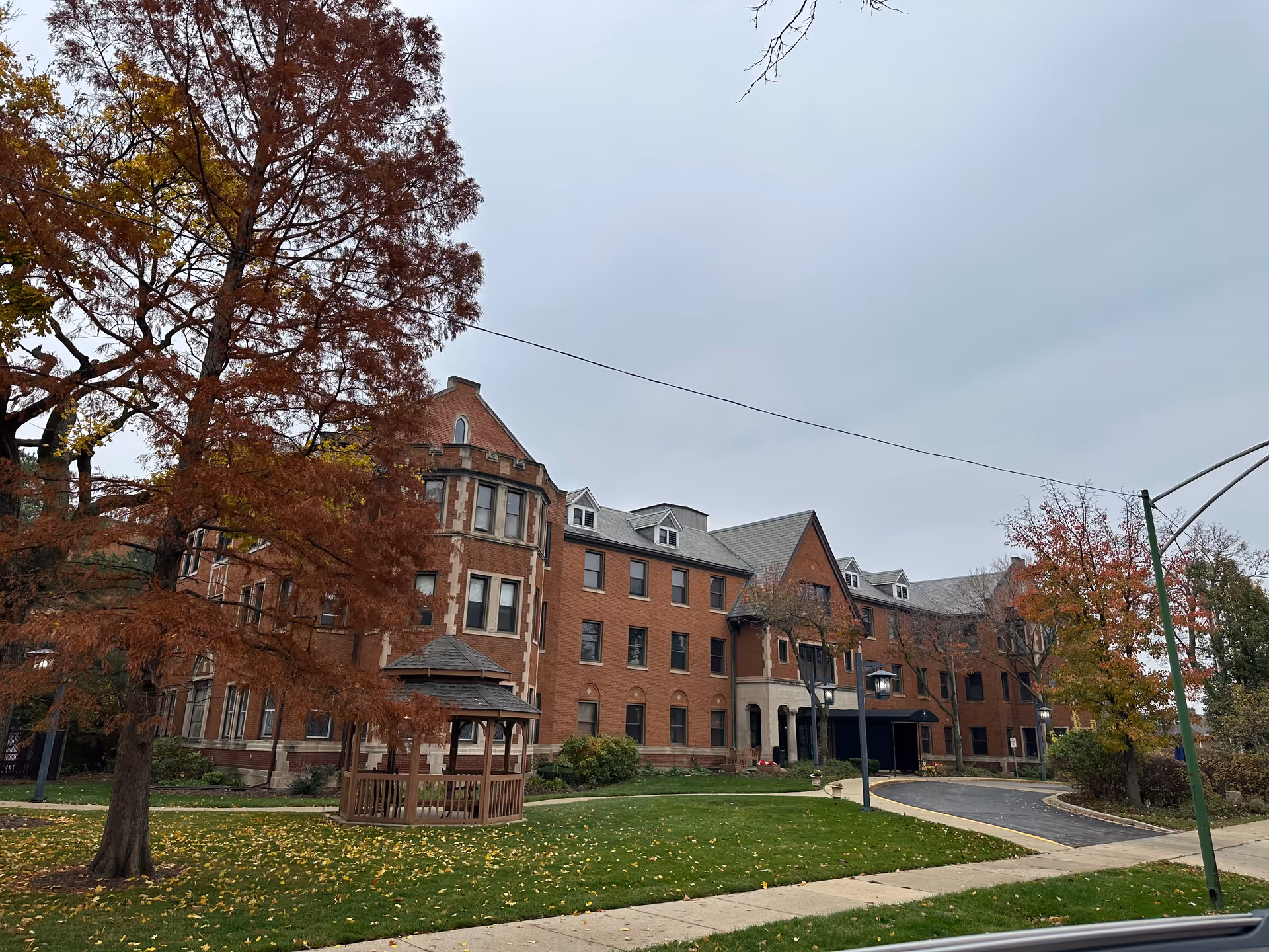 Brick multi-story senior living building with a small gazebo and autumn trees on a lawn under an overcast sky.