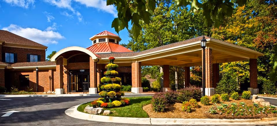 Front entrance of a brick retirement community building with a covered porte-cochere, landscaped roundabout, and tiered flower display.